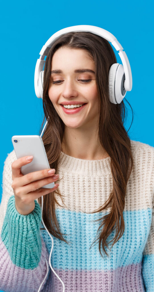 Technology, millennials and lifestyle concept. Carefree cute brunette female student put on headphones, plug-in smartphone picking song and smiling, standing blue background make playlist for study
